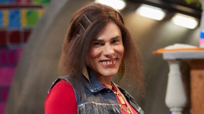 Graça (Rodrigo Sant'Anna) sorri com vestido vermelho, colete jeans e brincos grandes em cena da série Tô de Graça