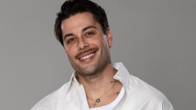 Gui Araújo sorri para a câmera com camisa branca aberta sobre camiseta clara diante de fundo cinza liso