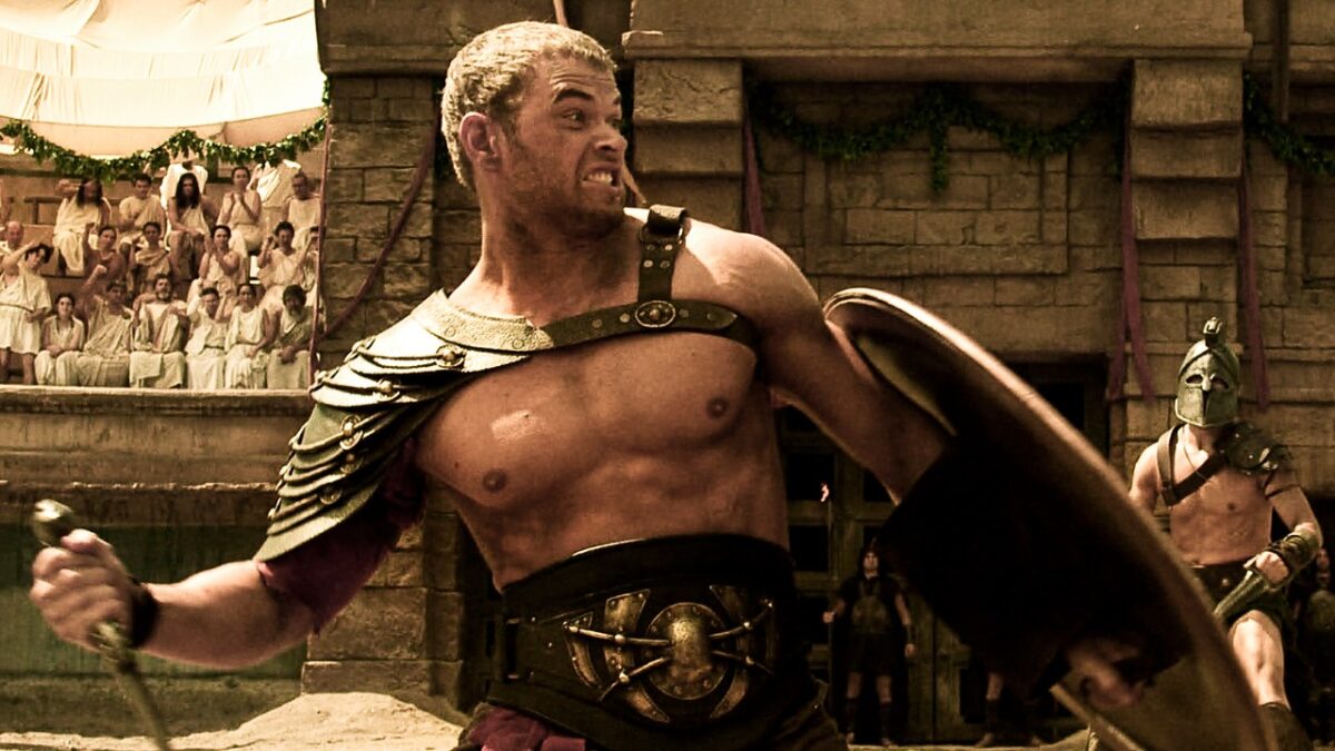 Hércules (Kellan Lutz) empunha espada e escudo em arena de combate com guerreiros e plateia ao fundo