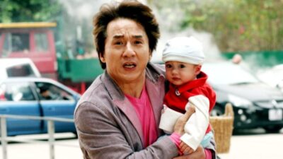 Jackie Chan segura Matthew Medvedev no colo com expressão de preocupação em rua com carros ao fundo em Três Ladrões e um Bebê