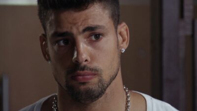 Jorginho (Cauã Reymond) com expressão séria, camiseta branca e corrente prateada em cena da novela Avenida Brasil