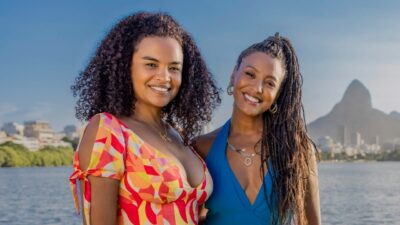 Juli (Lucy Ramos) e Bela (Sheron Menezzes) sorriem juntas à beira da lagoa, com montanhas do Rio ao fundo