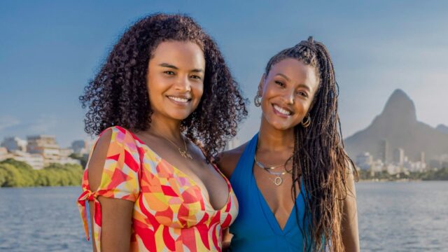 Juli (Lucy Ramos) e Bela (Sheron Menezzes) sorriem juntas à beira da lagoa, com montanhas do Rio ao fundo