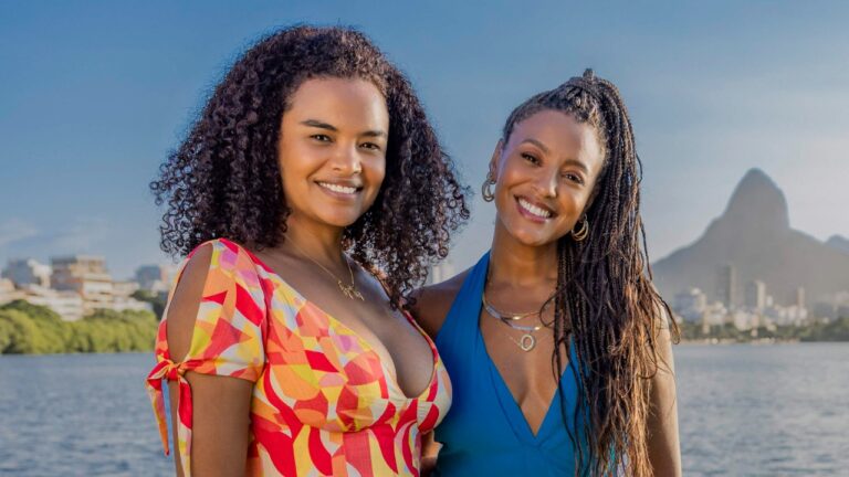 Juli (Lucy Ramos) e Bela (Sheron Menezzes) sorriem juntas à beira da lagoa, com montanhas do Rio ao fundo