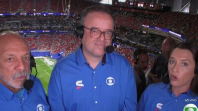 Junior, Everaldo Marques e Ana Thaís Matos de headset na cabine de transmissão com estádio e gramado ao fundo durante a Copa