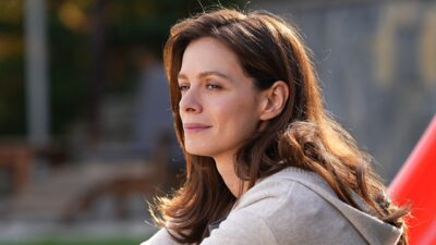 Karsu (Ozge Ozpirincci) com leve sorriso e olhar distante ao ar livre usando casaco claro em cena da novela Coração de Mãe