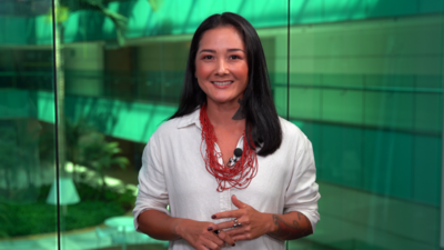 Mulher de cabelo preto e blusa branca sorrindo em frente a painel de vidro verde com microfone preso à roupa