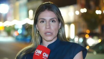 Ludmila Candal segura microfone da CNN Brasil durante reportagem em rua iluminada à noite com carros desfocados ao fundo