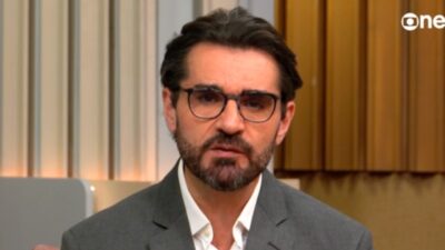Marcelo Cosme de blazer cinza e óculos fala diante da câmera no estúdio da GloboNews com painel de madeira ao fundo