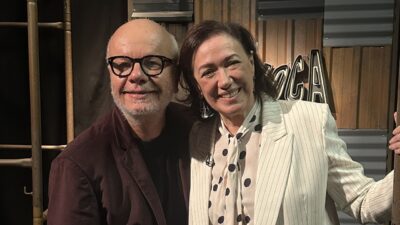 Marcelo Tas e Lilia Cabral sorriem abraçados no cenário do Provoca com painel de madeira e logotipo do programa ao fundo