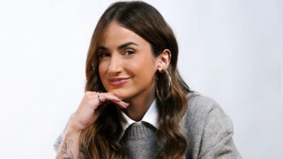 Mari Palma apoia o queixo na mão, sorri para a câmera e veste blusa cinza sobre camisa branca em fundo claro