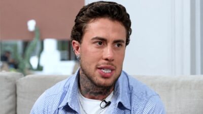 MC Daniel fala durante entrevista, com camisa listrada azul sobre camiseta branca, tatuagens no pescoço e sofá ao fundo