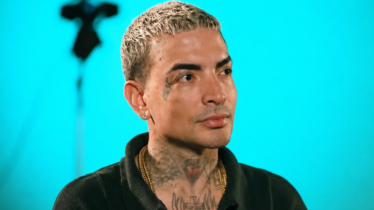 MC Guimê com expressão séria, cabelo platinado curto, tatuagens no rosto e pescoço durante entrevista em estúdio com fundo azul desfocado