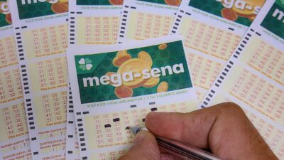 Mão marca números em volante da Mega-Sena com caneta sobre vários bilhetes das Loterias Caixa espalhados