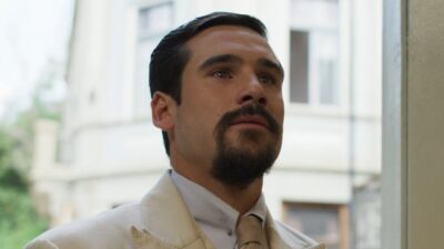 Mirinho (Nicolas Prattes) de terno claro com olhar atento diante de prédio em cena de A Nobreza do Amor