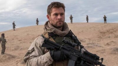Mitch Nelson (Chris Hemsworth) segura fuzil em área desértica com soldados ao fundo em cena de 12 Heróis