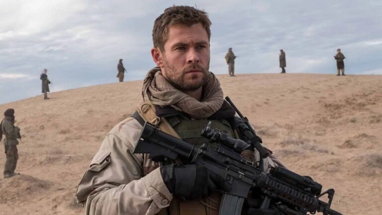 Mitch Nelson (Chris Hemsworth) segura fuzil em área desértica com soldados ao fundo em cena de 12 Heróis