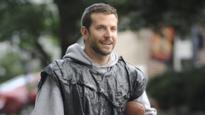 Pat (Bradley Cooper) sorri usando moletom cinza com capuz e saco plástico preto sobre os ombros enquanto segura uma bola de futebol americano ao ar livre em cena de O Lado Bom da Vida