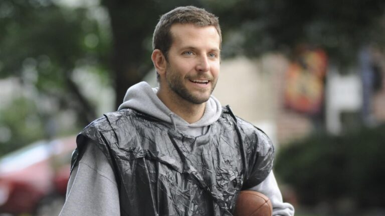 Pat (Bradley Cooper) sorri usando moletom cinza com capuz e saco plástico preto sobre os ombros enquanto segura uma bola de futebol americano ao ar livre em cena de O Lado Bom da Vida
