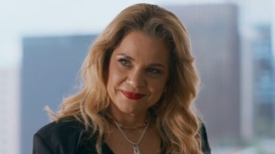 Pilar (Isabel Teixeira) sorri com batom vermelho, blazer preto e colares em cena da novela Quem Ama Cuida com ambiente interno desfocado ao fundo