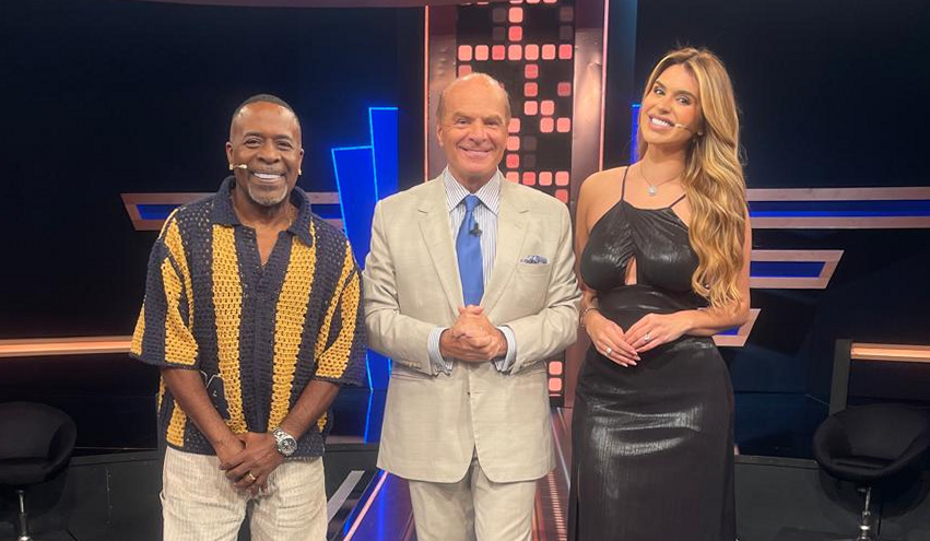 Pimpolho, Marcelo de Carvalho e Mônica Fonseca sorriem lado a lado no palco do Mega Senha com painel iluminado ao fundo