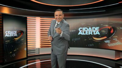 Reinaldo Gottino de terno cinza aponta para a câmera no estúdio do Cidade Alerta com telões do programa ao fundo