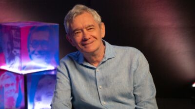 Serginho Groisman de camisa azul-clara sorri para a câmera diante de cubos iluminados com retratos em fundo escuro