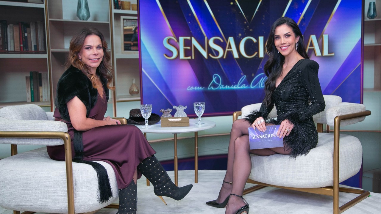 Sula Miranda e Daniela Albuquerque sentadas frente a frente em poltronas durante entrevista no Sensacional com mesa ao centro e telão do programa ao fundo
