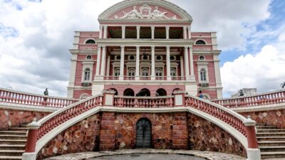 Fachada do Teatro Amazonas em tons de rosa e branco com colunas, escadarias simétricas e céu nublado ao fundo