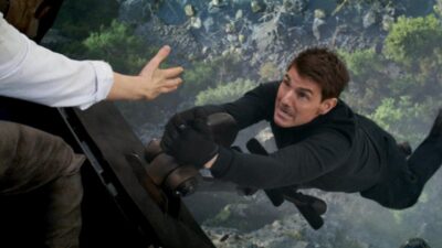 Ethan Hunt (Tom Cruise) pendurado na lateral de um helicóptero estende a mão para outra pessoa sobre um desfiladeiro em cena de Missão Impossível: Acerto de Contas Parte 1