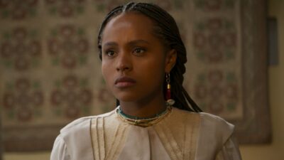 Alika/Lúcia (Duda Santos) de tranças, roupa clara e colares olha séria à frente em cena de A Nobreza do Amor