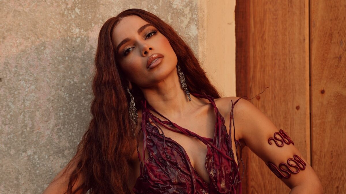 Anitta de cabelo ruivo longo posa com vestido vinho e adornos nos braços diante de parede e porta de madeira