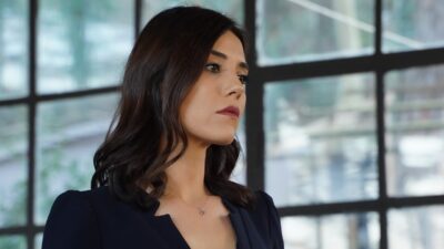Asya (Cansu Dere) de cabelo escuro e blazer preto olha para o lado diante de grandes janelas em cena de Iludida