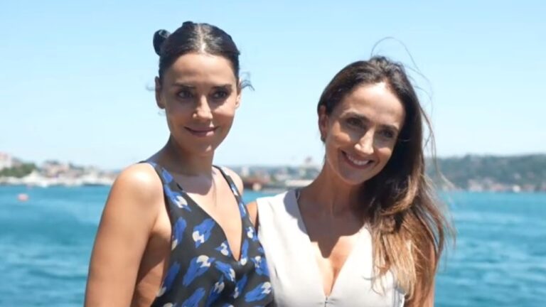 Cansu Dere e Patrícia Nielsen sorrindo lado a lado diante do mar em dia ensolarado