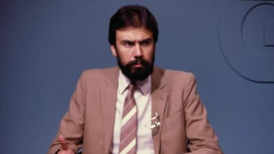 Carlos Monforte de terno bege e gravata listrada, com barba, sentado e gesticulando diante de fundo azul