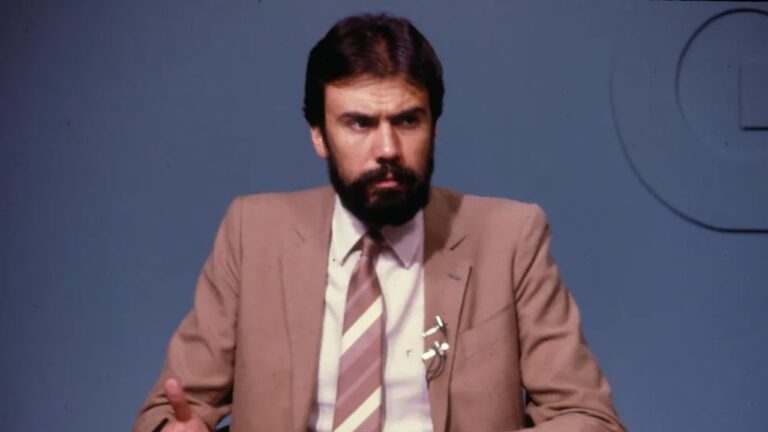 Carlos Monforte de terno bege e gravata listrada, com barba, sentado e gesticulando diante de fundo azul