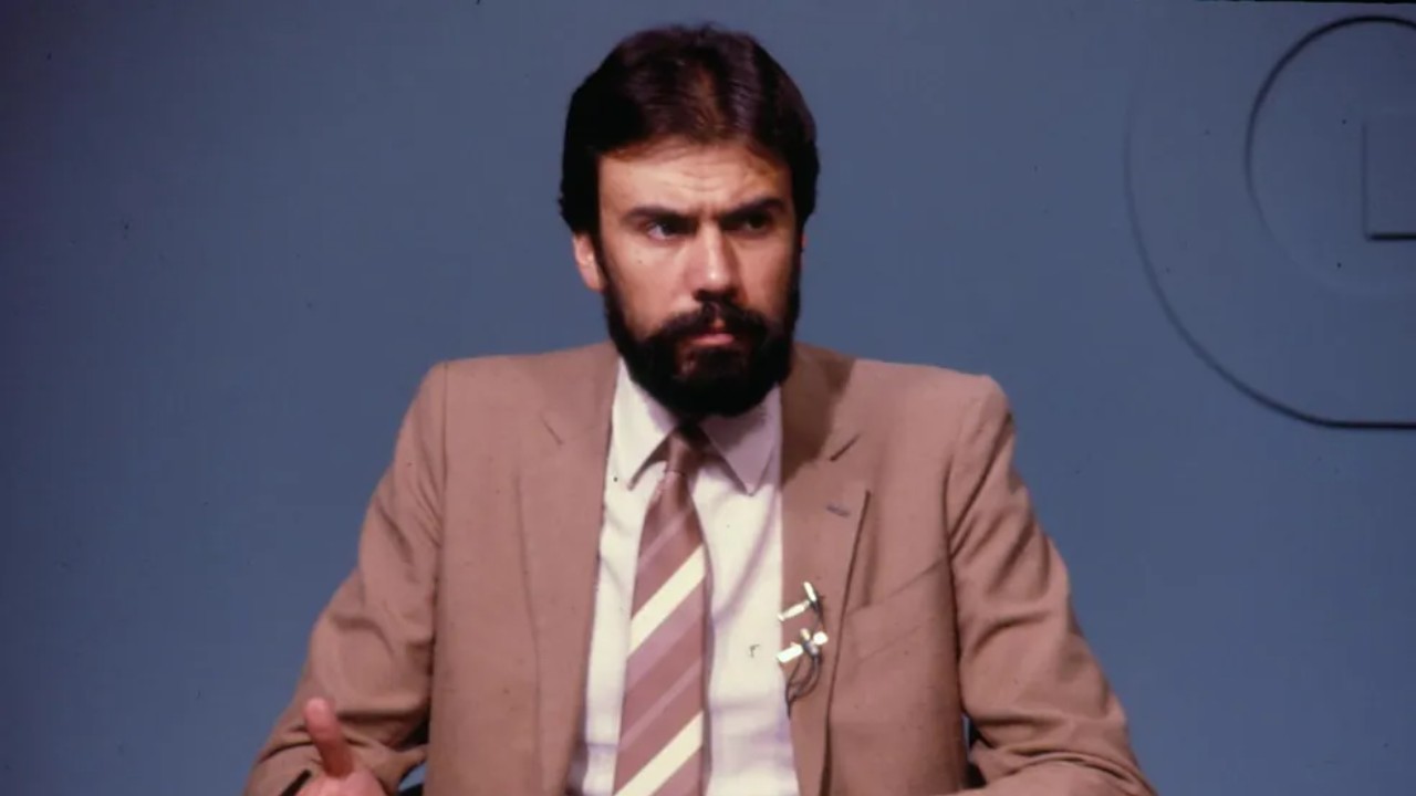 Carlos Monforte de terno bege e gravata listrada, com barba, sentado e gesticulando diante de fundo azul