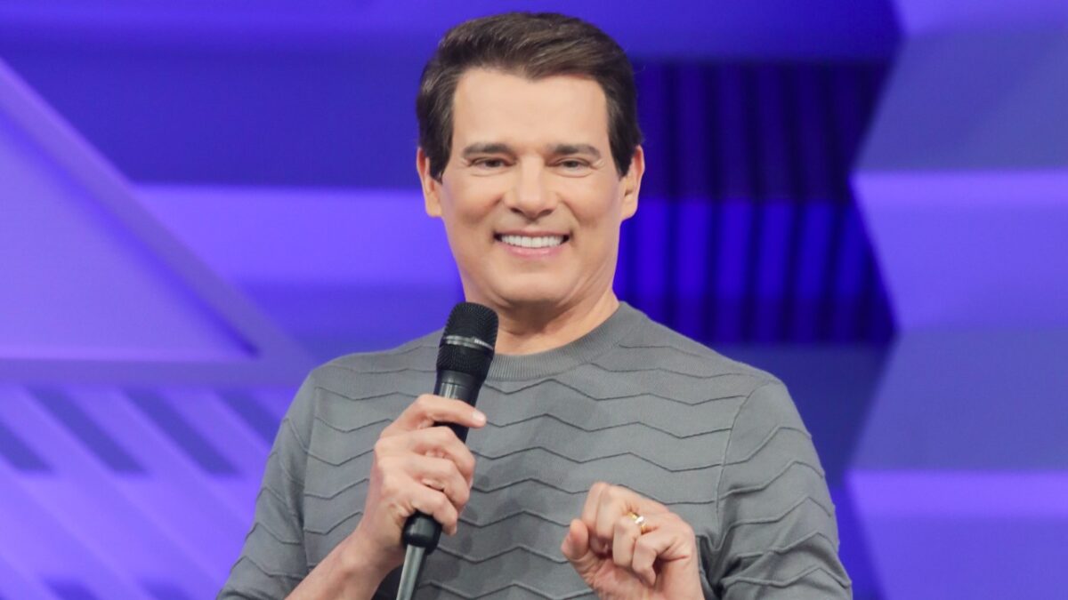 Celso Portiolli sorrindo de blusa cinza segura microfone diante de telão roxo em estúdio de TV