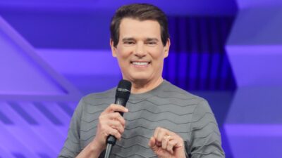 Celso Portiolli sorrindo de blusa cinza segura microfone diante de telão roxo em estúdio de TV