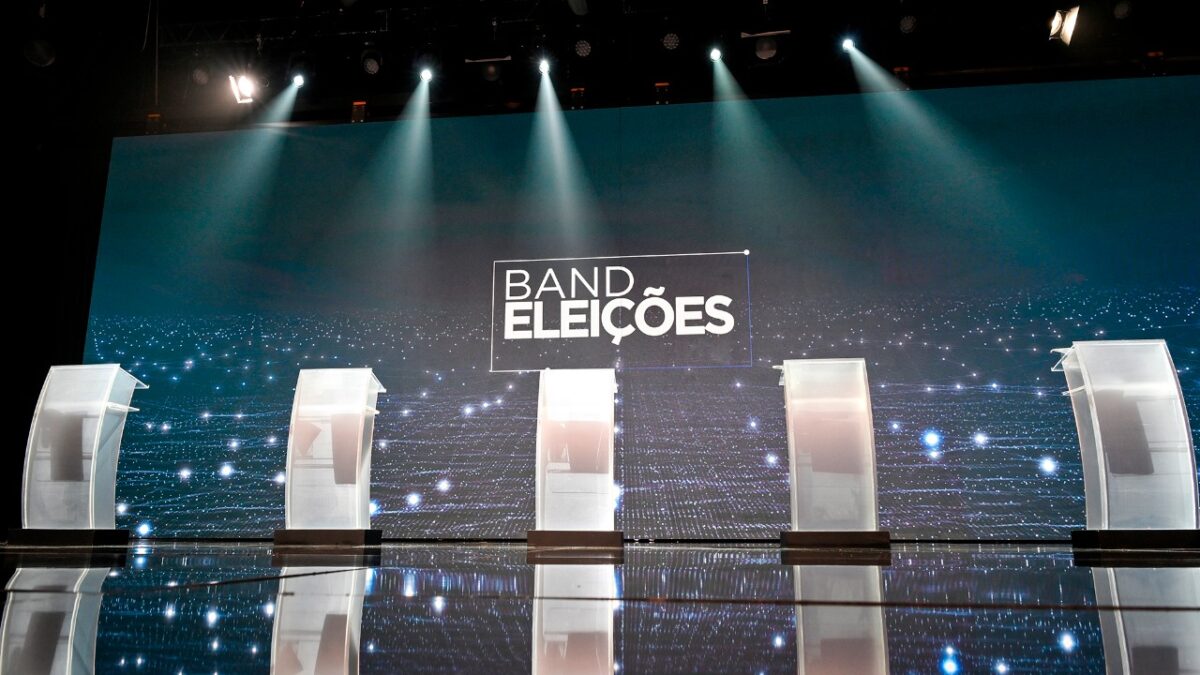 Cenário do debate da Band com cinco púlpitos, telão Band Eleições ao fundo e refletores sobre o palco