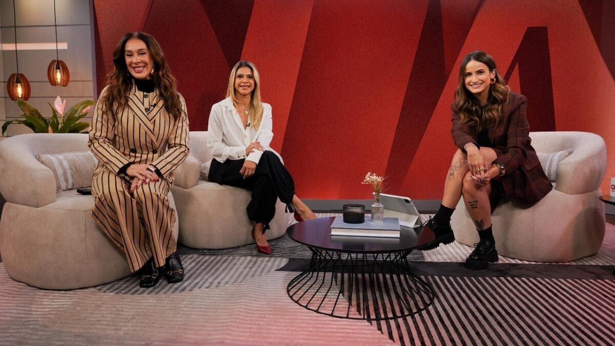 Claudia Raia, Sylvana Vasconcelos e Mari Palma sentadas e sorrindo em estúdio com sofás e painel vermelho ao fundo
