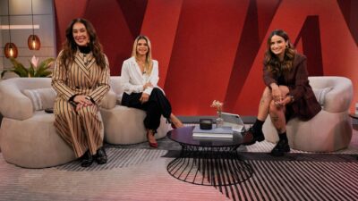 Claudia Raia, Sylvana Vasconcelos e Mari Palma sentadas e sorrindo em estúdio com sofás e painel vermelho ao fundo