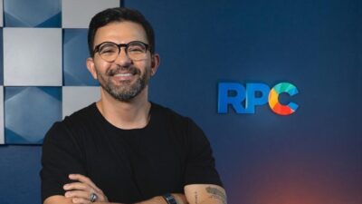 Duani Reis de camiseta preta sorri de braços cruzados diante de parede azul com logotipo da RPC
