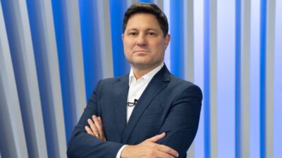 Eleandro Passaia de terno azul e camisa branca posa de braços cruzados em estúdio com painel azul ao fundo