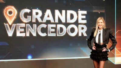 Gianne Albertoni sorri de blazer e saia pretos diante de telão com o logotipo do programa O Grande Vencedor