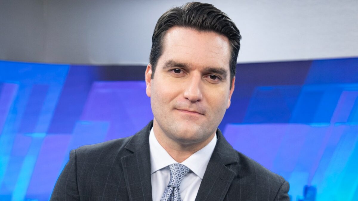 Gustavo Toledo de terno e gravata olha para a câmera diante de painel azul no estúdio da Record News