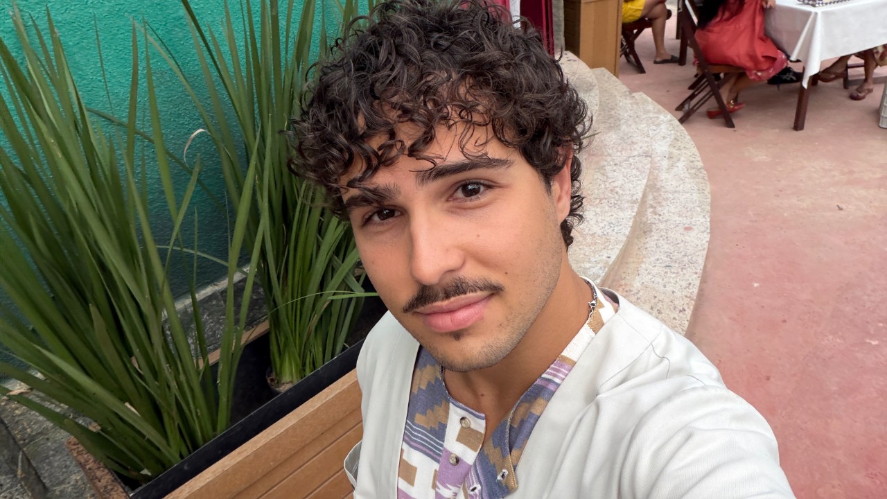 Igor Jansen faz selfie de camisa clara, com cabelo cacheado e bigode, em área externa com plantas e mesas ao fundo