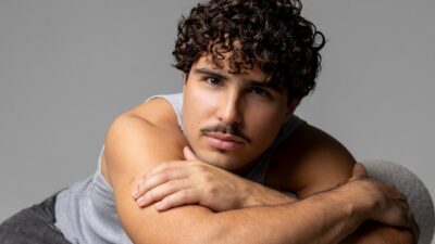 Igor Jansen de regata cinza posa com os braços cruzados, cabelo cacheado e bigode, diante de fundo cinza
