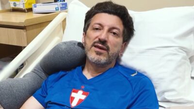 Lito Sousa de camisa azul com símbolo da Suíça, deitado em cama de hospital com travesseiro branco