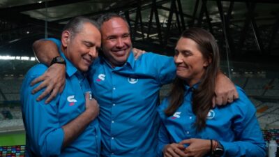 Luis Roberto, Caio Ribeiro e Ana Thaís Matos de camisas azuis se abraçam e sorriem em cabine de estádio